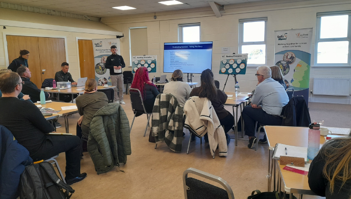 A meeting room with people sat at round tables.  There is a man stood at the front of the room presenting with a screen showing the text Telling the Story