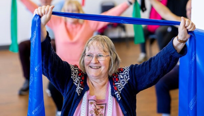 an older lady taking part in a seated exercise class. She is holding a blue exercise band above her head