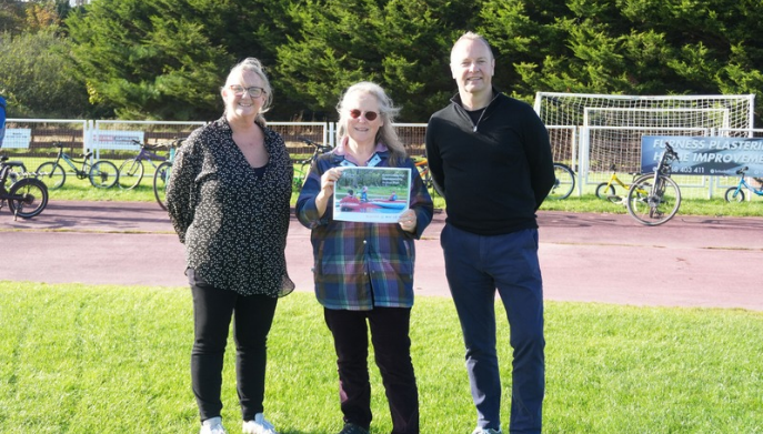 Three people stand on a grass field beside an outdoor cycle track. The person in the centre is holding a document. Several bicycles are visible in the background alongside a football goal and advertising boards, with trees lining the edge of the venue on a bright, sunny day.